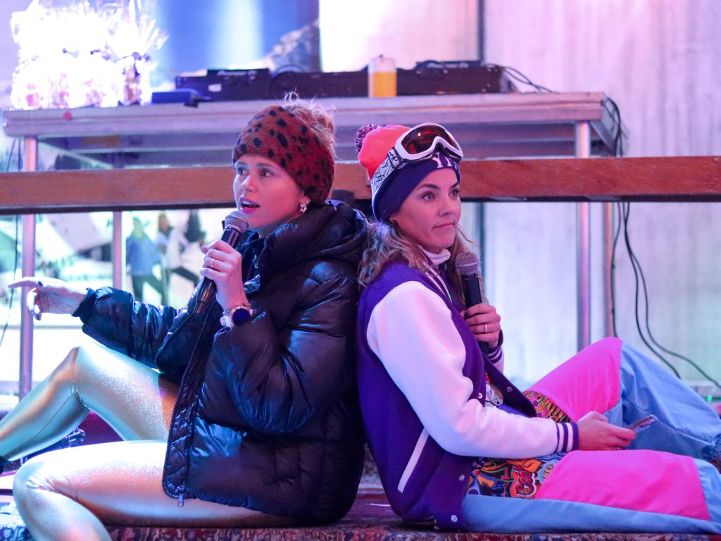 Een foto van Heleen en Eva die op het podium zitten in skikleding en een speech geven.