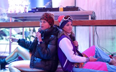 Een foto van Heleen en Eva die op het podium zitten in skikleding en een speech geven.
